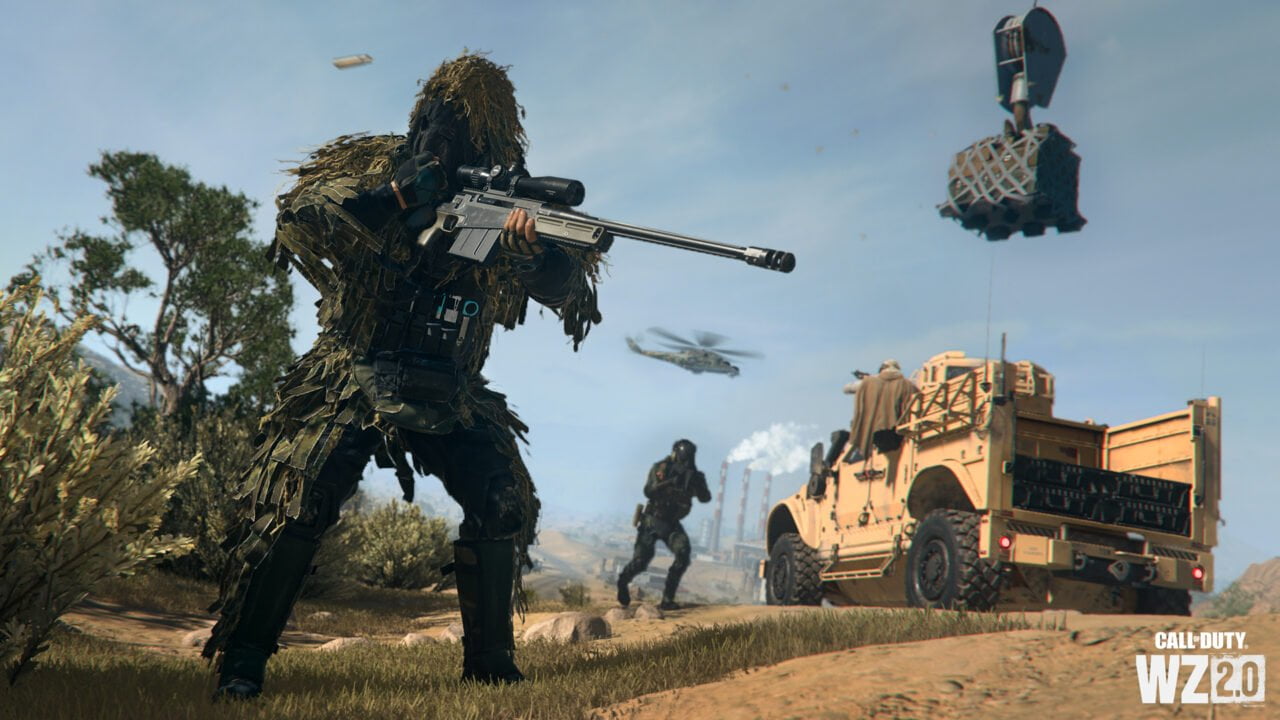 Call of Duty: Warzone Mobile. Postać w stroju maskującym z karabinem snajperskim na pierwszym planie z pojazdem wojskowym i helikopterem w tle na pustynnym krajobrazie, widoczne logo "Call of Duty WZ 2.0".