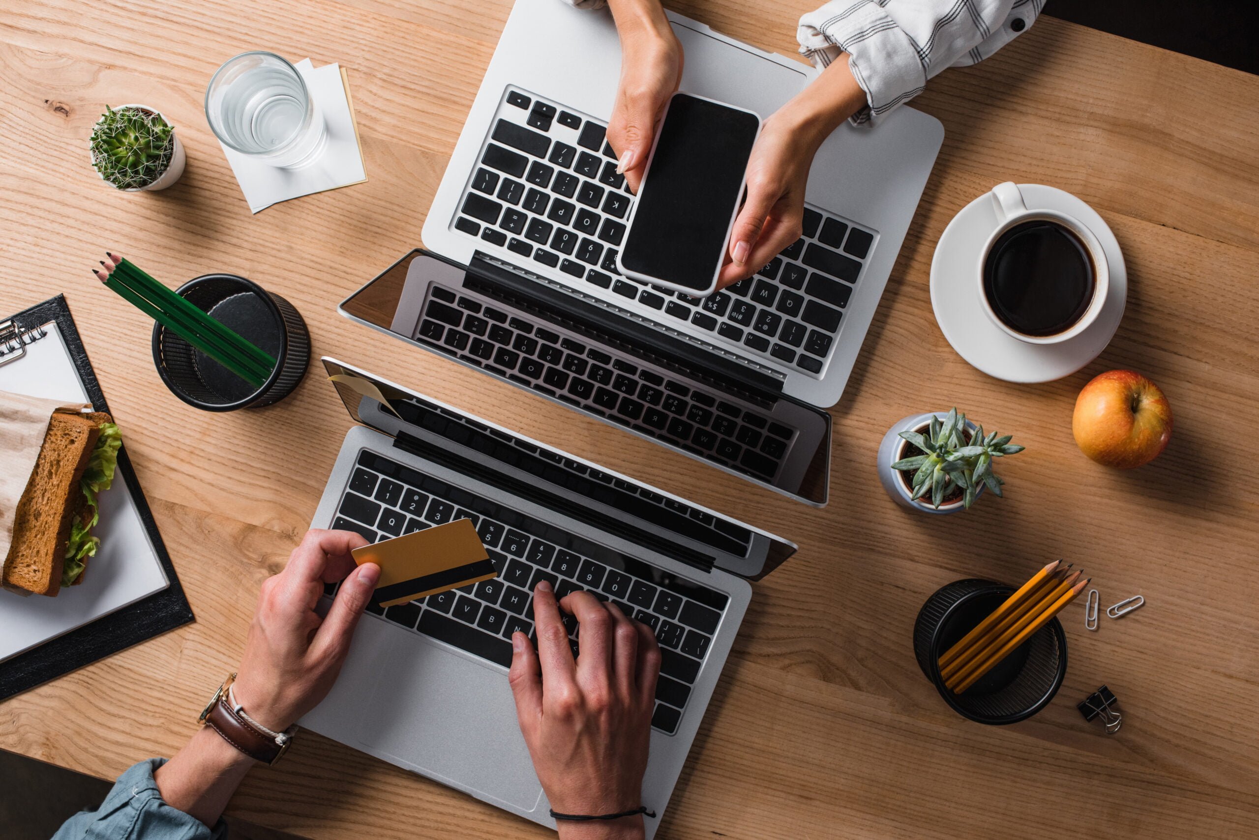 Dwie osoby pracują przy biurku z laptopami, jedna wkłada kartę kredytową do slotu na laptopie, a druga trzyma smartfona nad klawiaturą; na biurku kawa, kanapka, jabłko i artykuły biurowe.