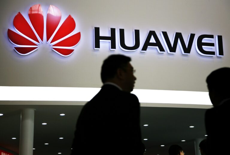 Silhouette dwóch mężczyzn przed podświetlonym logo firmy Huawei na wystawie.