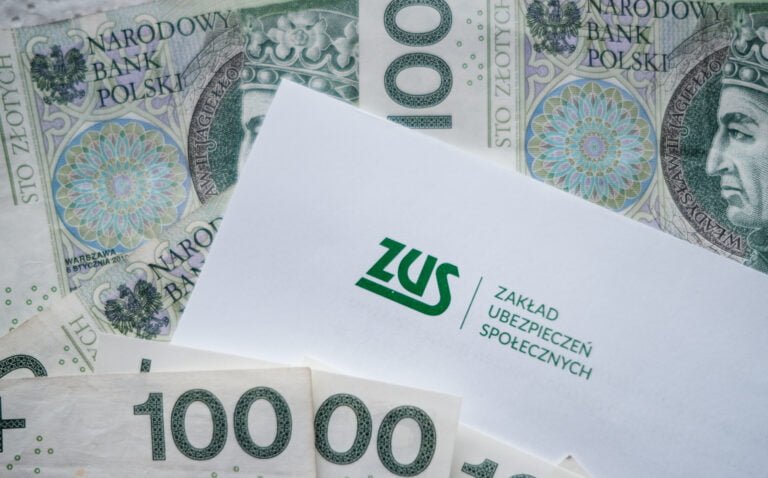 Zdjęcie koperty z logo ZUS na pierwszym planie i banknotów 100-złotowych w tle.