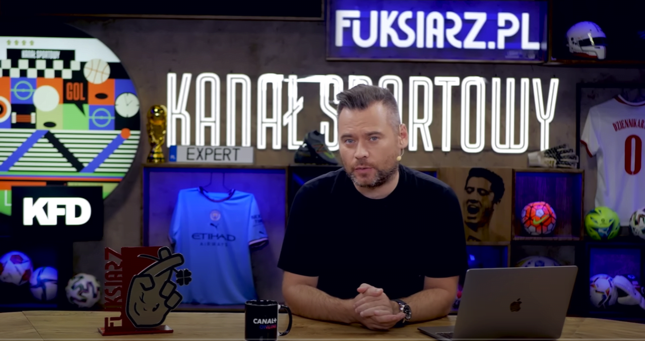 Mężczyzna siedzący za biurkiem w studiu z gadżetami sportowymi w tle, w tym koszulkami piłkarskimi i piłkami, a nad nim neonowy napis "KANAŁ SPORTOWY" i logo "FUKSIARZ.PL".