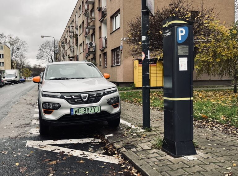 Biały samochód marki Dacia zaparkowany przy automacie do opłat parkingowych na tle bloku mieszkalnego, ulica zarośnięta drzewami w okresie jesieni, pochmurne niebo.