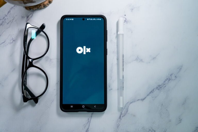 Smartfon z logo OLX na ekranie, położony na marmurowym blacie obok okularów i białego długopisu.