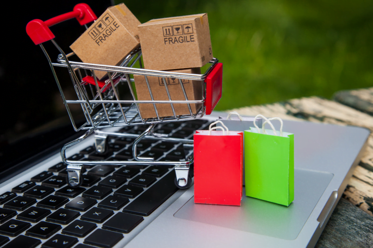 Mały wózek sklepowy z kartonowymi pudełkami z napisem "FRAGILE" stoi na laptopie, obok dwie małe kolorowe torby na zakupy online
