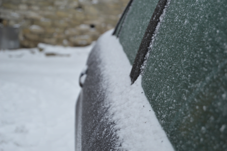 Samochód pokryty śniegiem i szronem, w tle niewyraźny widok na kamienne konstrukcje.