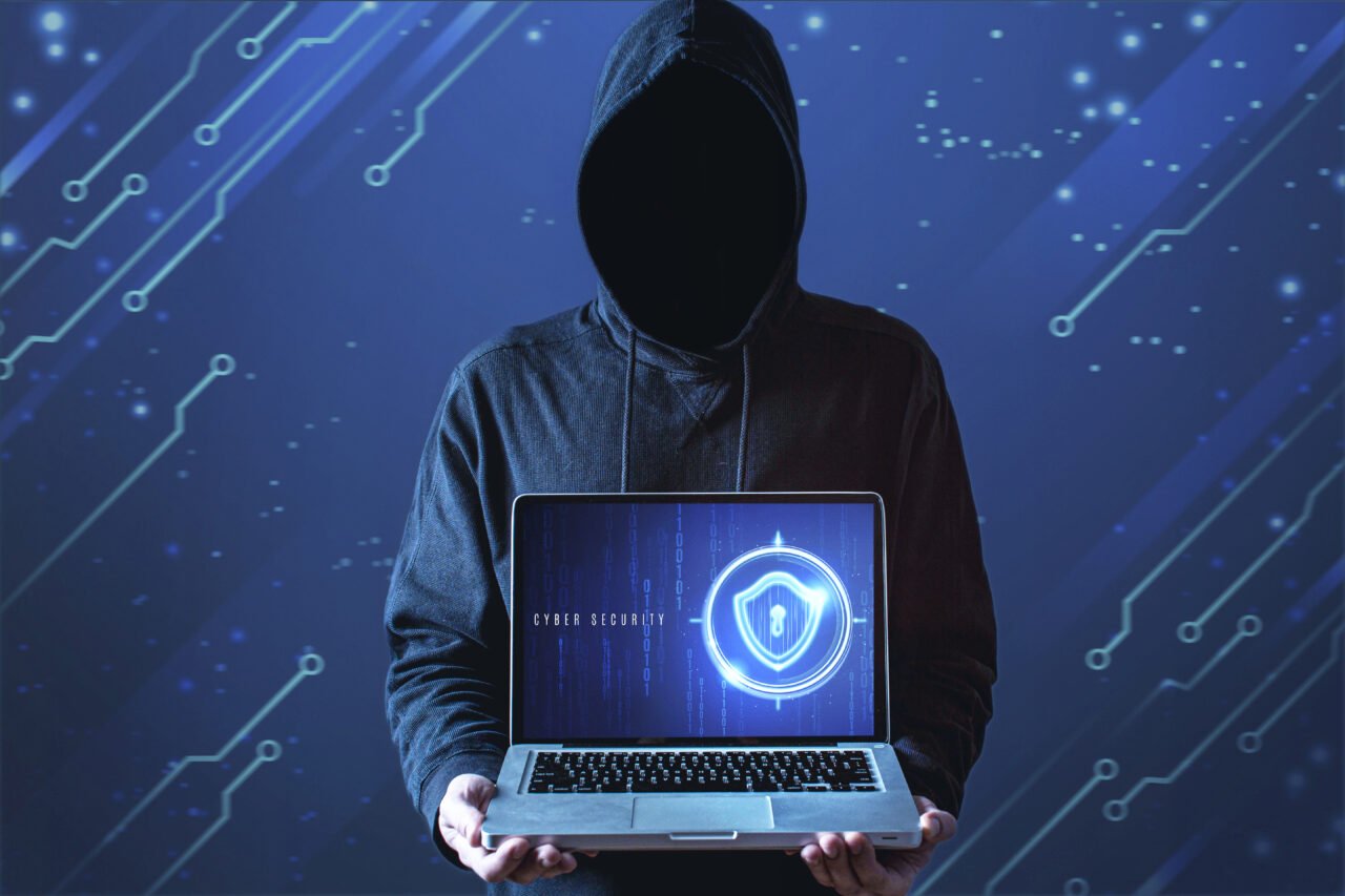 Osoba w kapturze trzymająca laptopa z wyświetlonym symbolem cyberbezpieczeństwa na tle cyfrowych obwodów. programy antywirusowe