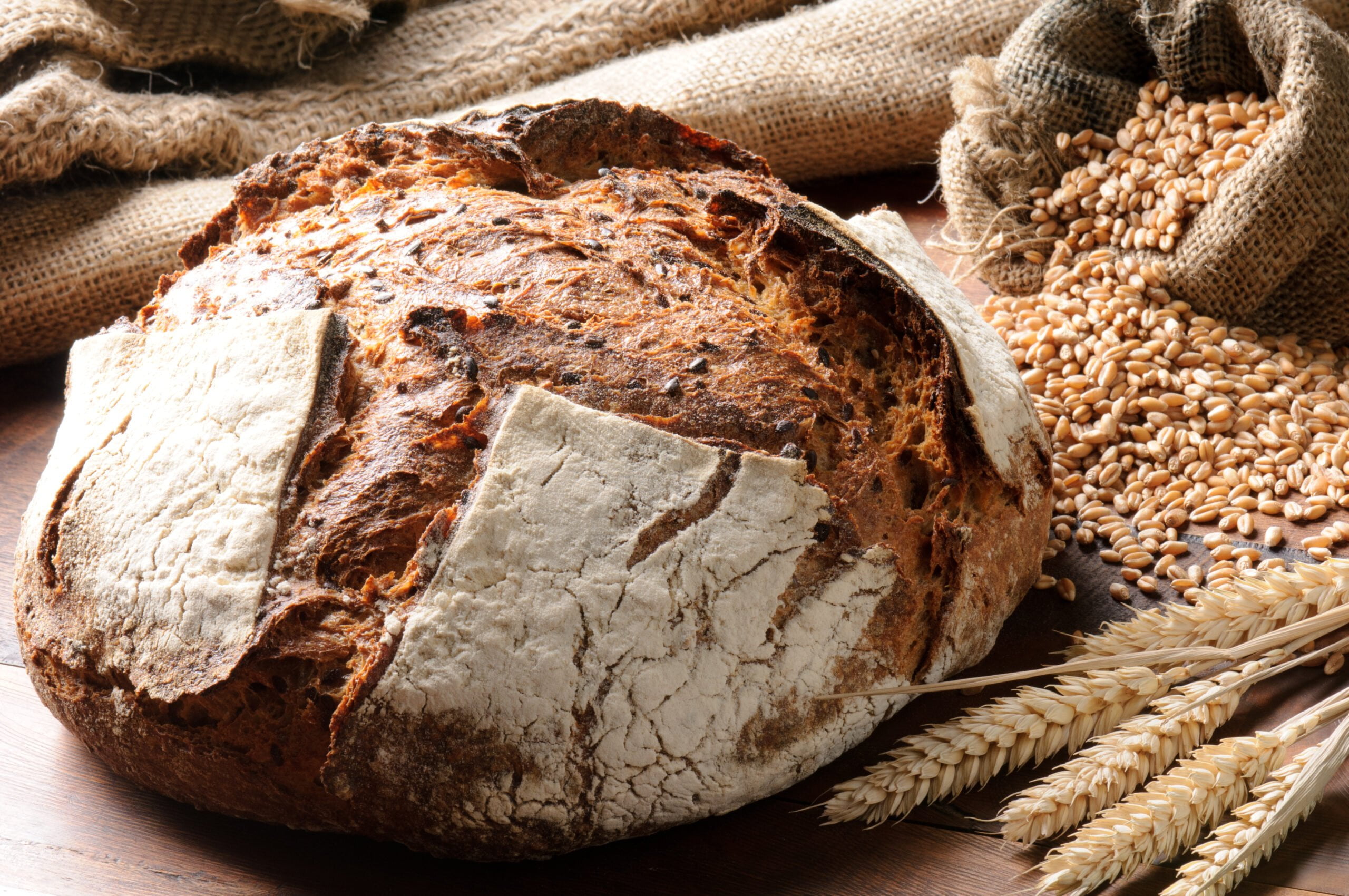 Duży bochenek chleba na drewnianym blacie, obok worka pełnego ziaren pszenicy i kłosów pszenicy na pierwszym planie.