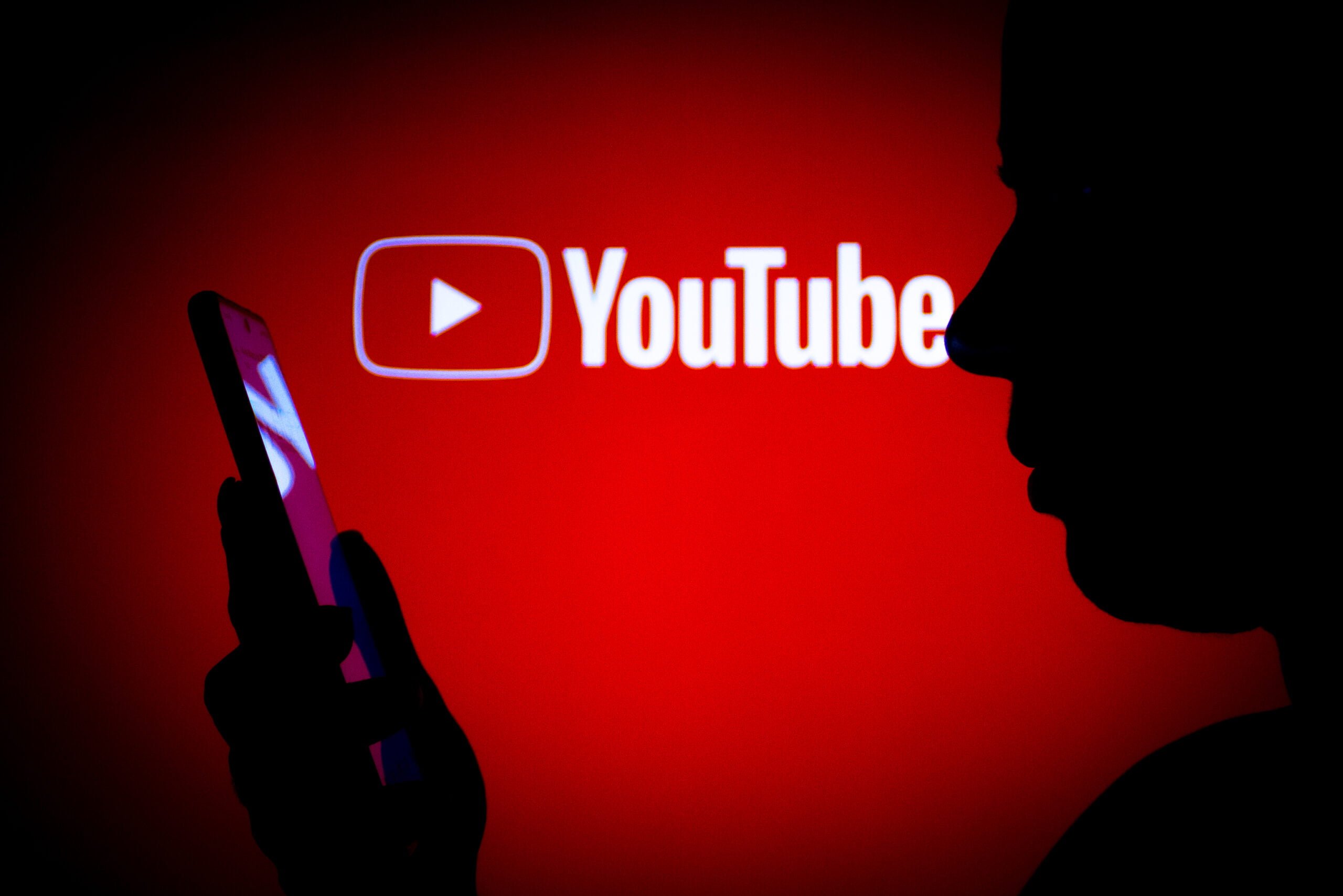 Sylwetka osoby trzymającej smartfon na czerwonym tle z logo YouTube.