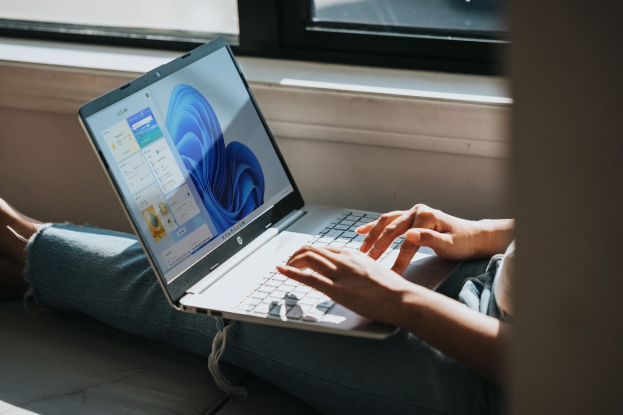 Osoba siedząca przy oknie z laptopem z Windows 11 na kolanach, pracująca na klawiaturze; na ekranie laptopa pulpit systemu Windows, na którym w przyszłości może się znaleźć model phi-3-mini
