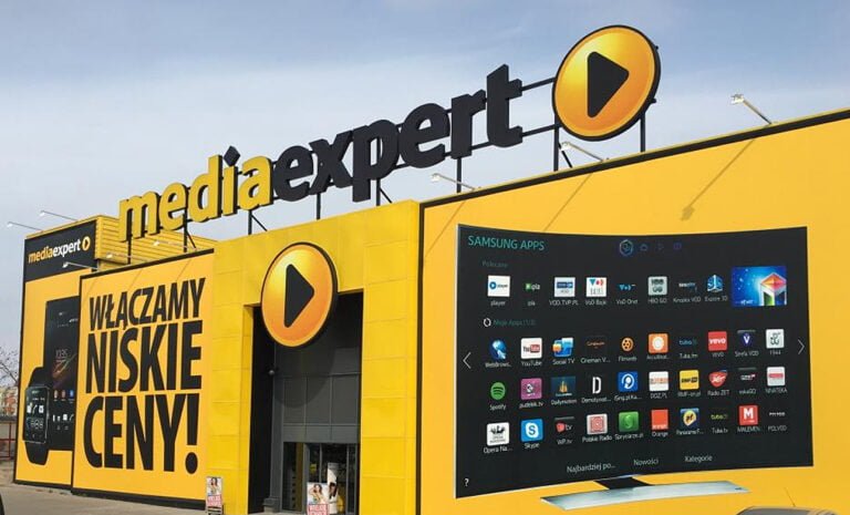 Fasada sklepu Media Expert w kolorze żółtym z dużym logo i napisem "Włączamy niskie ceny!". Obok, duży ekran z wyświetlanymi aplikacjami.