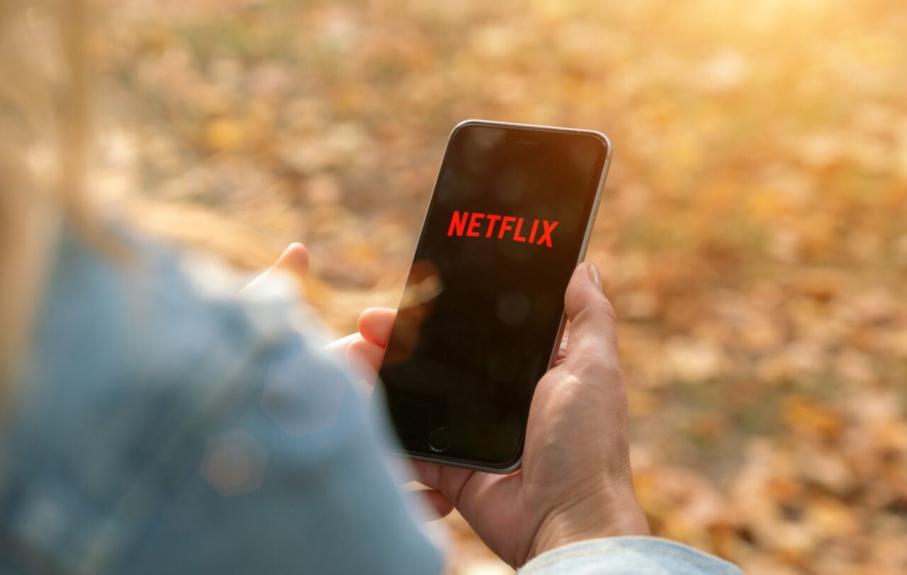 Osoba trzymająca smartfon z logo Netflix na ekranie na tle rozmytych jesiennych liści.