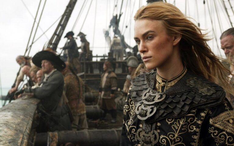 Kobieta w zdobionej zbroi stoi na pokładzie statku, za nią załoga w epoce żeglarskiej.