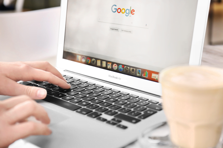 Osoba używająca laptopa MacBook Air z otwartą stroną główną Google UK na ekranie, obok stoi kubek kawy latte. Jak wylogować się z konta google?