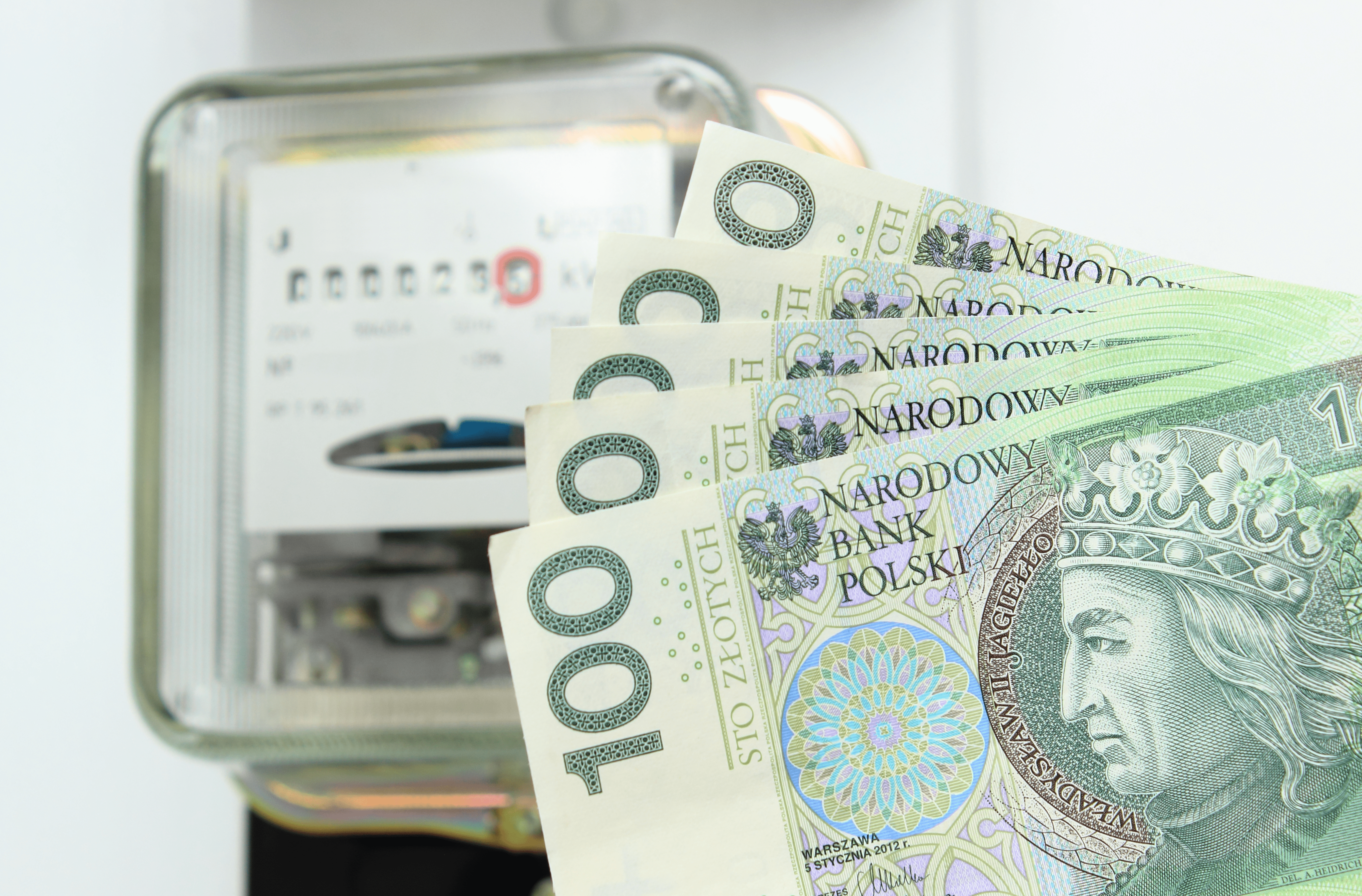 Sto złotych banknoty z wizerunkiem króla Władysława Jagiełły przed rozmazanym tłem z elektrycznym licznikiem zużycia energii. Fotowoltaika w 2024 roku pozwoli dużo zaoszczedzić za rachunki za prąd