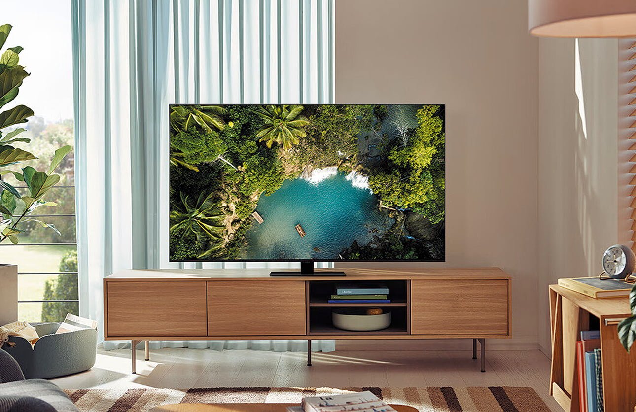 Wnętrze salonu z dużym telewizorem Samsunga na drewnianej komodzie wyświetlającym zdjęcie tropikalnych krajobrazów; w tle widok przez okno i dekoracje w pokoju.
