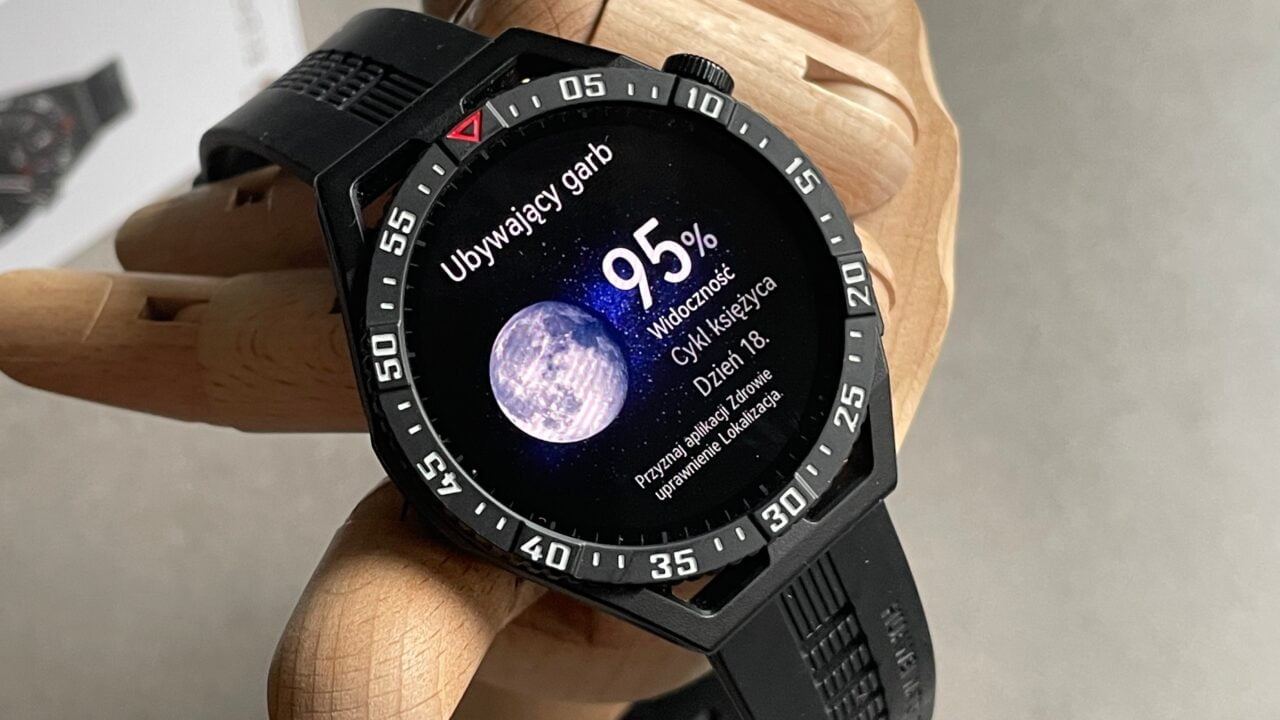 Cyfrowy zegarek sportowy na drewnianej ręce manekina wyświetlający obraz księżyca i informacje o jego aktualnej fazie.