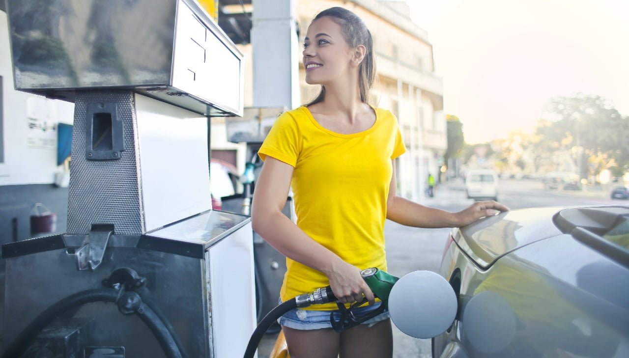 Młoda kobieta w żółtej koszulce tankuje paliwo E10 do samochodu na stacji benzynowej.