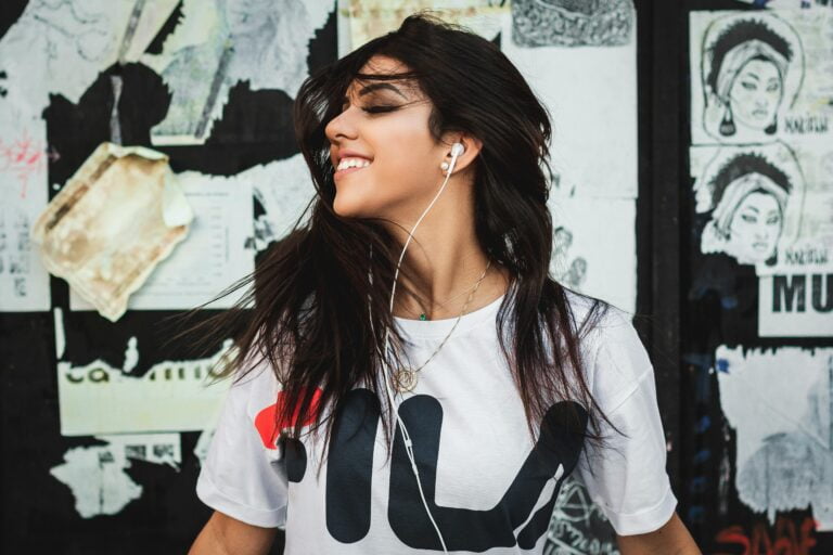 Młoda kobieta z zamkniętymi oczami i uśmiechem na twarzy tańczy, słuchając muzyki na słuchawkach. Ma na sobie białą koszulkę z logo. Tło stanowią miejskie plakaty i graffiti.