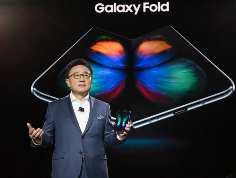 Mężczyzna w garniturze prezentuje smartfon na tle grafiki przedstawiającej rozłożony telefon Galaxy Fold.