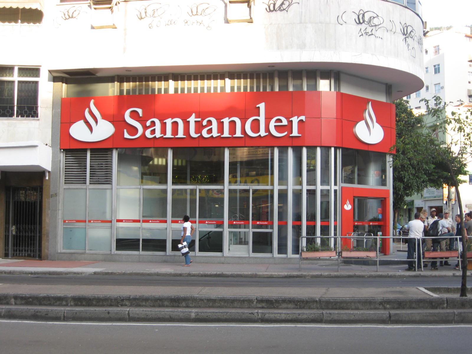 Oddział banku Santander z czerwoną tablicą i logo, przed którym ludzie rozmawiają, a jeden przechodzi obok na chodniku.