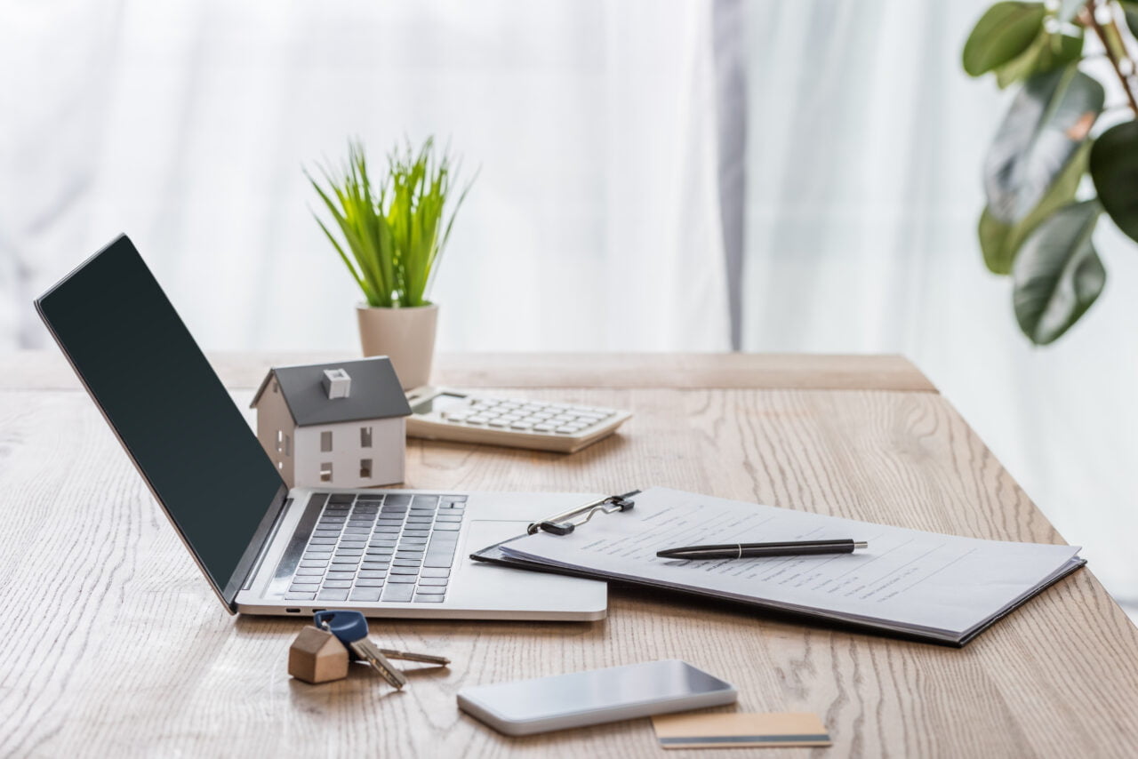 Przykładowe biurko z otwartym laptopem, modelowym domkiem, kluczami, doniczką z rośliną, kalkulatorem, notatnikiem i długopisem na drewnianym stole.