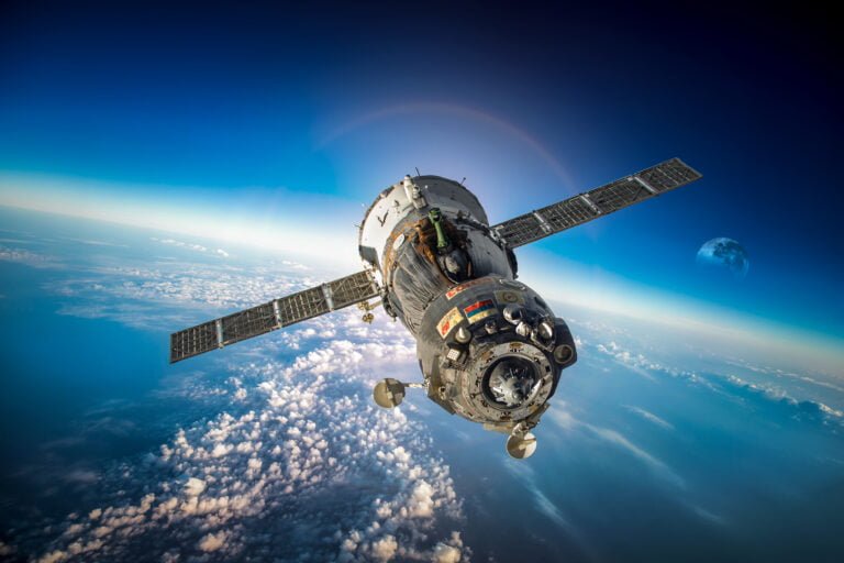 Statek kosmiczny Sojuz na orbicie okołoziemskiej z widocznymi panelami słonecznymi, Ziemią i częściowo widocznym Księżycem w tle.