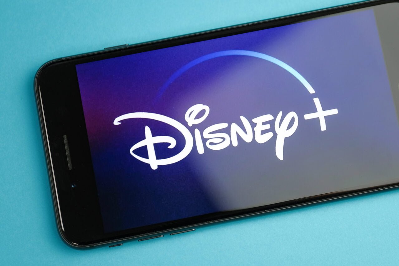 Logo Disney+ wyświetlone na smartfonie, który leży na niebieskim tle. Grafika do artkykułu "Disney+ cena. Ile kosztuje Disney+ w 2023 roku?"