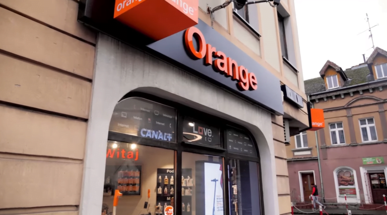 Widok na front sklepu Orange z logo firmy, wystawami sklepowymi i napisem "Witaj" na szybie.