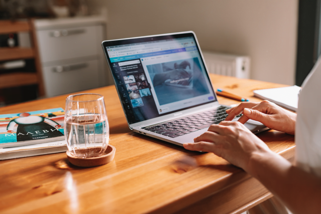 Praca zdalna Osoba korzystająca z laptopa na drewnianym biurku w domowym biurze, na ekranie widoczna strona internetowa z grafiką rekina, obok laptopa szklanka wody i magazyn.