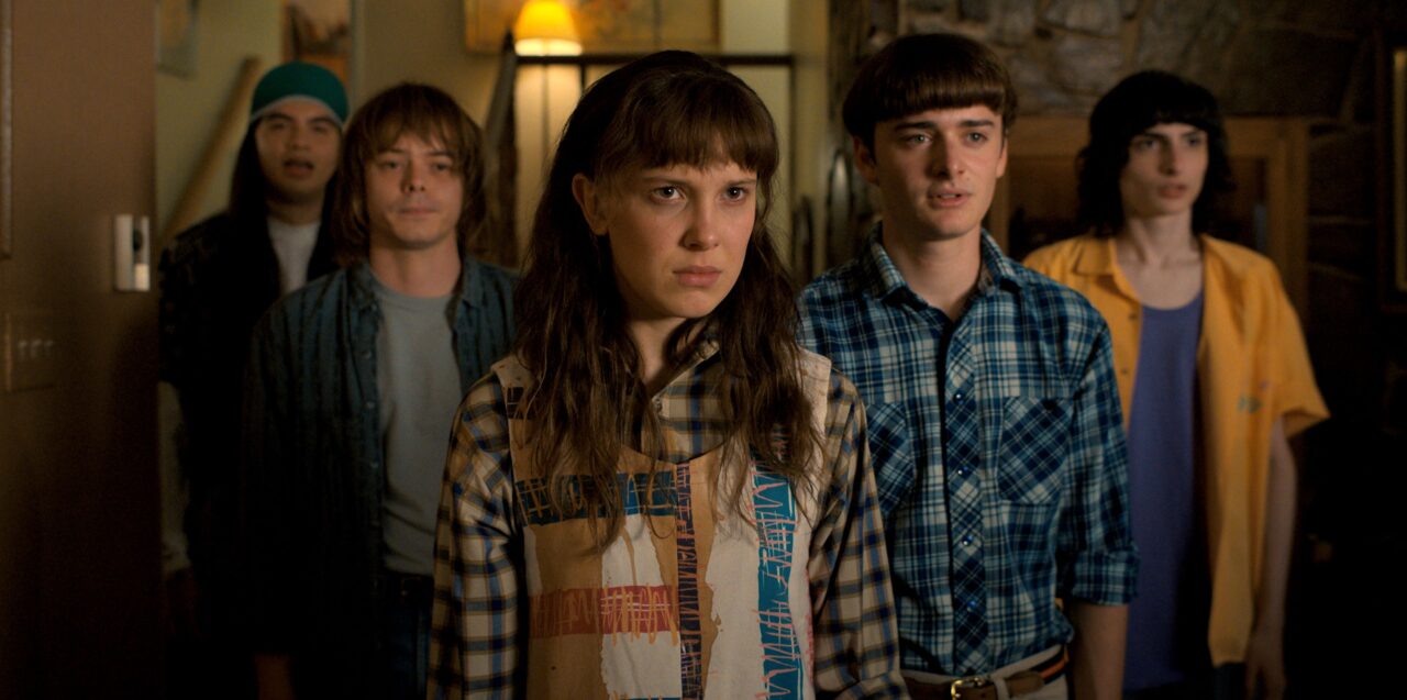 Kadr z serialu Stranger Things od Netflixa. Piątka nastoletnich przyjaciół stoi w pokoju, patrząc poważnie przed siebie; na pierwszym planie dziewczyna z długimi brązowymi włosami i grzywką.