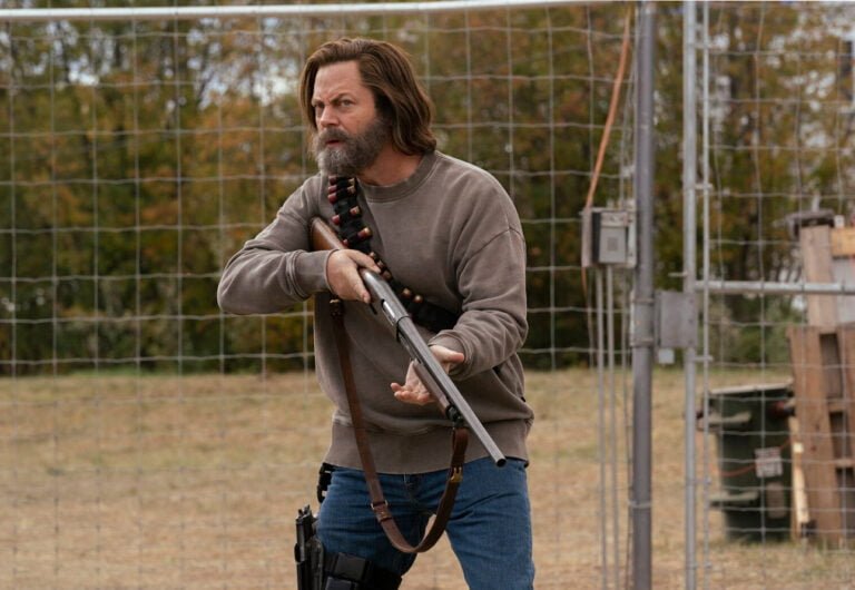 Mężczyzna z brodą trzymający strzelbę z amunicją przewieszoną przez ramię, stojący przed ogrodzeniem z siatki. Kadr z serialu The Last of Us od HBO