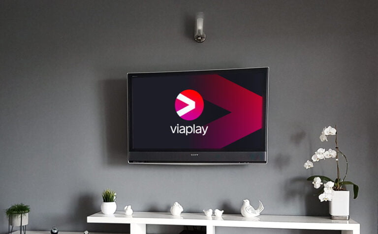 Telewizor na ścianie wyświetlający logo serwisu Viaplay, umieszczony nad białą komodą z dekoracyjnymi przedmiotami i doniczką z kwiatami.