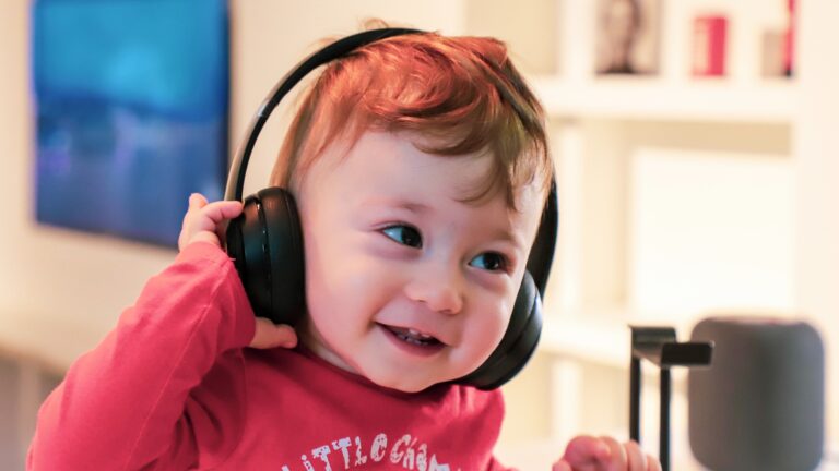 Uśmiechnięte dziecko w czerwonej bluzie trzymające na głowie słuchawki na tle rozmytego wnętrza.