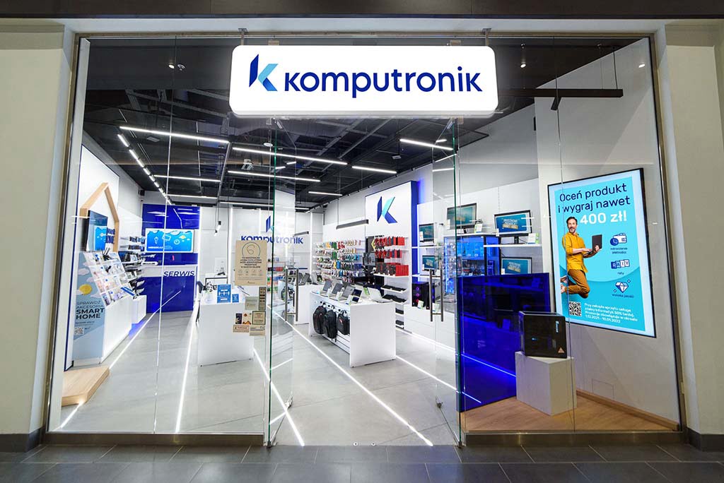Wnętrze sklepu Komputronik z produktami elektronicznymi na półkach, ekspozycyjnymi ekranami i reklamami oraz logotypem firmy nad wejściem.