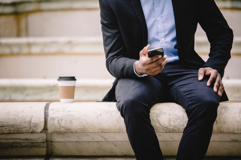 Mężczyzna w garniturze siedzi na kamiennym murku z telefonem komórkowym w ręce i kartonowym kubkiem kawy obok siębie.