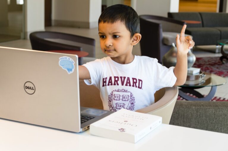 Mały chłopiec w białej koszulce z napisem "HARVARD" siedzi przy biurku i podnosi palec w górę, patrząc na laptop dla dzieci