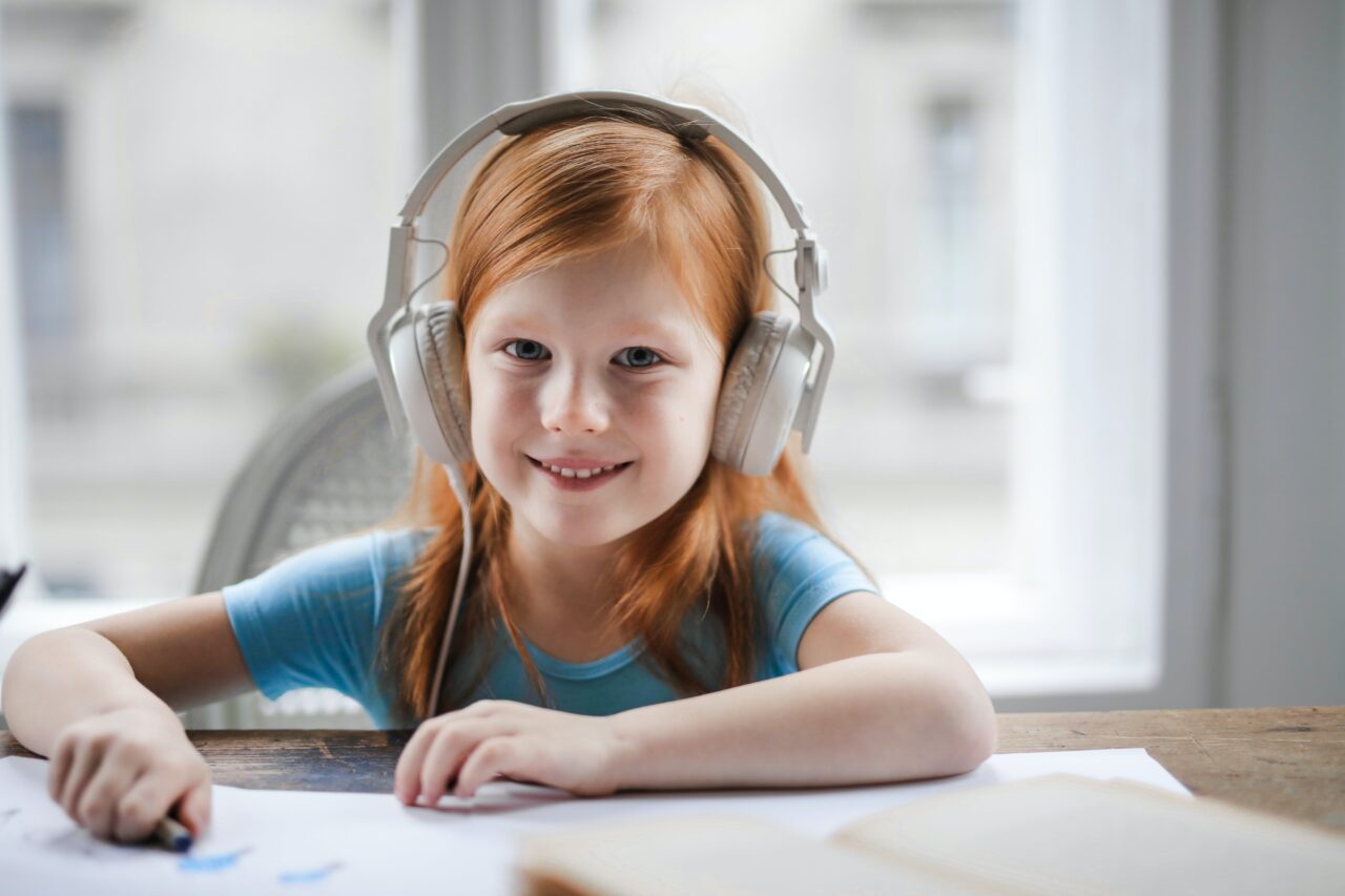 Ruda dziewczynka z uśmiechem na twarzy nosi słuchawki nauszne i rysuje coś na kartce papieru, przy stole z otwartą książką.