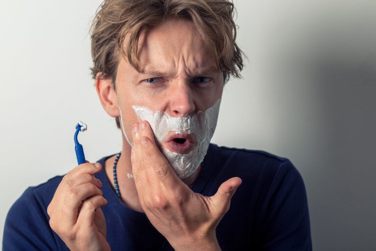 Mężczyzna z zaniepokojoną miną trzyma maszynkę do golenia i dotyka twarzy z pianką do golenia, prawdopodobnie sprawdzając skaleczenie po goleniu.