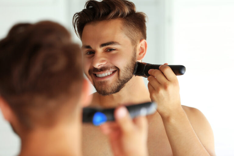 Młody mężczyzna przycina brodę elektryczną maszynką do golenia, oglądając się w lustrze.