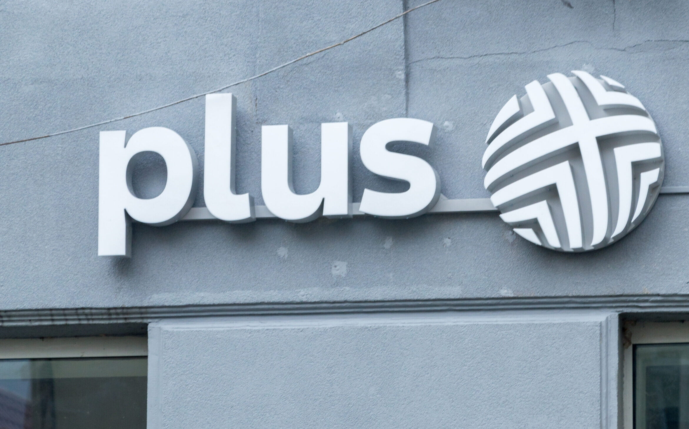 Widok na logo "plus" zamocowane na ścianie zewnętrznej budynku, składające się z białych liter i kołowego, stylizowanego symbolu na szarym tle.