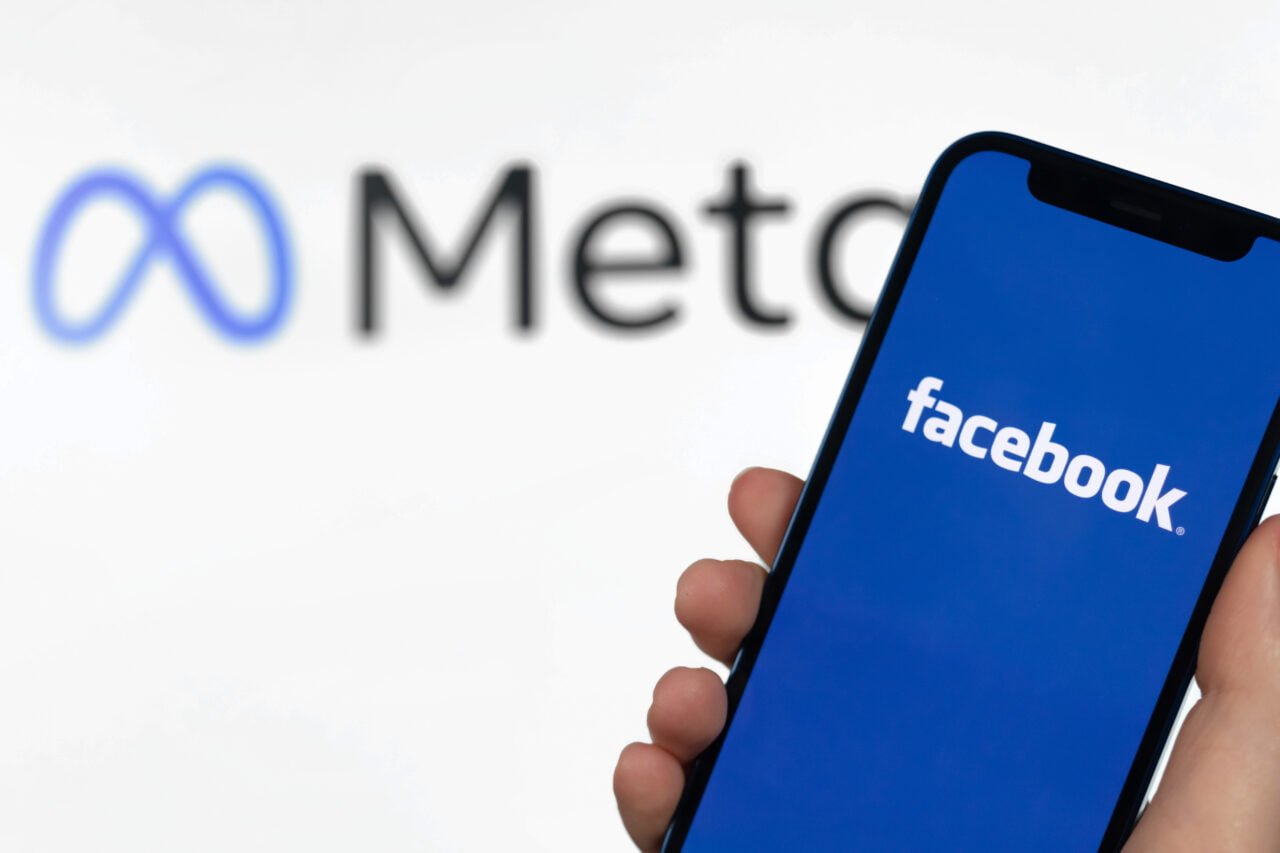 Oszustwo w mediach społęcznościowych. Dłoń trzymająca smartfon z logo Facebooka na ekranie, w tle rozmyte logo firmy Meta.