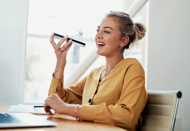 Kobieta w żółtej koszuli używa telefonu jako głośnomówiącego podczas rozmowy, siedząc przy biurku i uśmiecha się.