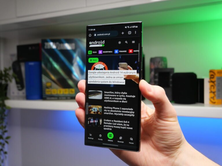 Ręka trzymająca smartfon Galaxy S23 Ultra z otwartą stroną internetową o tematyce technologicznej na tle rozmytego biurka z komputerowym sprzętem w pomieszczeniu z zielonym oświetleniem.