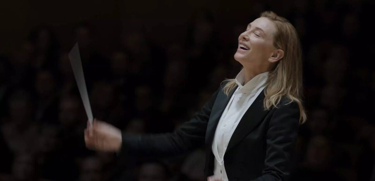 Cate Blanchett
w filmie Tar. Dyrygentka orkiestry w czarnym garniturze z batutą uniesioną w powietrzu, wyrażająca radość podczas dyrygowania.