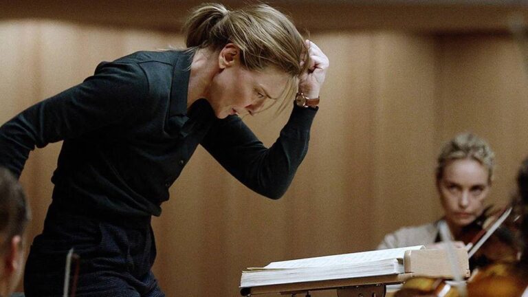 Cate Blanchett w filmie Tar. Dyrygentka orkiestry w ciemnym garniturze z zaciśniętą dłonią przy głowie skupia się na prowadzeniu muzyków, z nutami na pulpicie w tle.