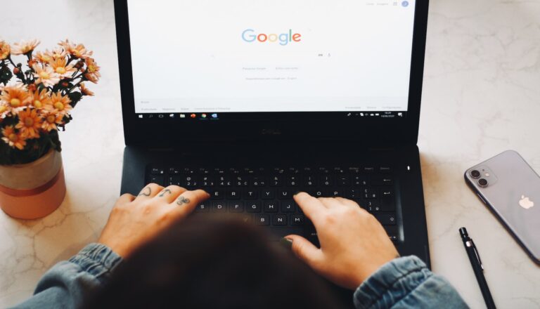 Osoba korzystająca z laptopa, na ekranie którego wyświetlona jest strona główna Google, obok komputera jest doniczka z kwiatami i telefon komórkowy.