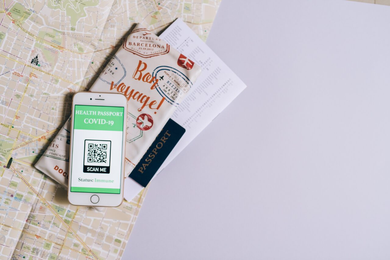 Smartfon z wyświetlonym certyfikatem zdrowotnym COVID-19 leży na mapie z widocznymi dokumentami podróży i paszportem.