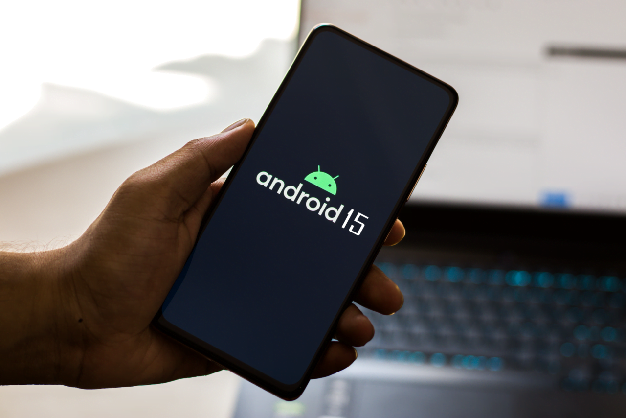 Debiut wersji Android 15 beta 1. Dłoń trzymająca smartfon z wyświetlonym logo systemu operacyjnego "Android 15" na ekranie, w tle niewyraźne elementy biurowe oraz klawiatura laptopa.
