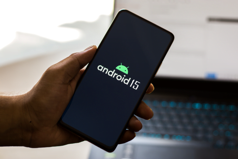 Dłoń trzymająca smartfon z wyświetlonym logo systemu operacyjnego "Android 15" na ekranie, w tle niewyraźne elementy biurowe oraz klawiatura laptopa.