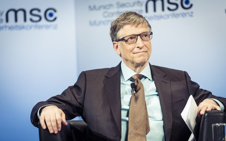 Bill Gates siedzi na konferencji, w tle banner z napisem "MSC Munich Security Conference", trzyma dokumenty i nosi okulary.